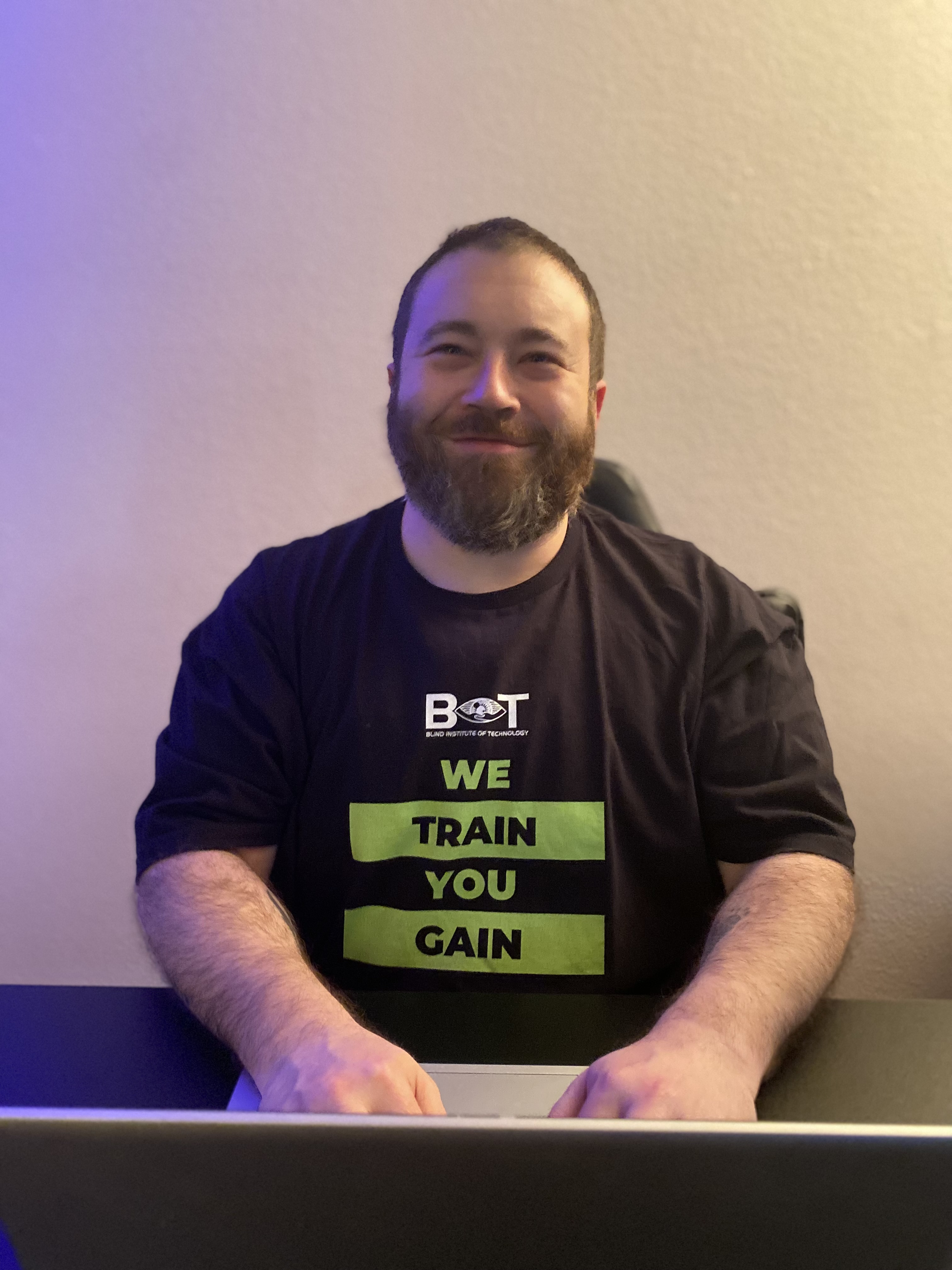 Jacques sitting at his desk, wearing his BIT T-shirt obtained while in his apprenticeship. The shirt is black with gold lettering.